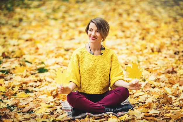 Niña sonriente en otoño sentada en la hierba y sosteniendo dos hojas amarillas en las manos