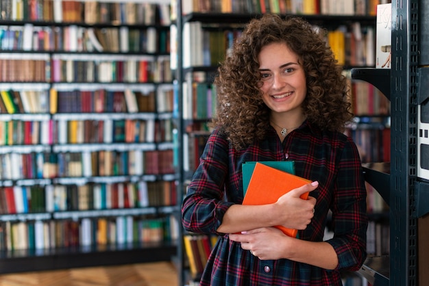 Niña sonriente de pie en la biblioteca abrazando libros en sus manos