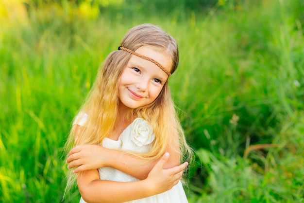 Niña sonriente en un prado verde, una niña pequeña se abraza a sí misma con un vestido blanco