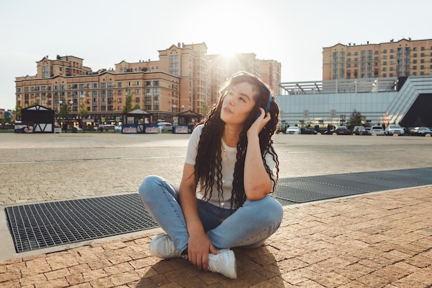 Una niña sonriente con rastas en los auriculares está sentada en el suelo en el parque Una joven feliz relajándose con auriculares disfrutando de la música Espacio para copiar la generación z