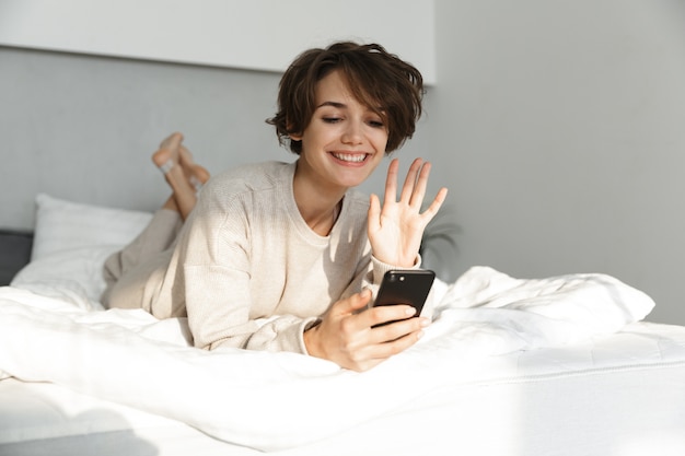 Niña sonriente relajándose en la cama por la mañana, tomando un selfie