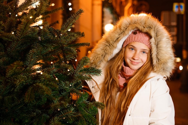 Niña sonriente en ropa de invierno con capucha celebra la navidad en la calle junto a cristo decorado ...