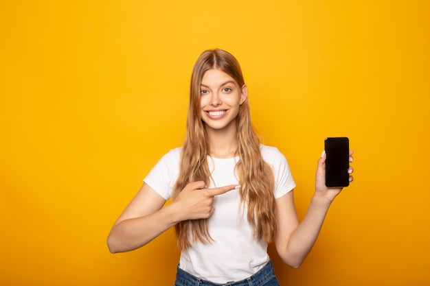 niña sonriente señalando con el dedo en el teléfono inteligente aislado en amarillo