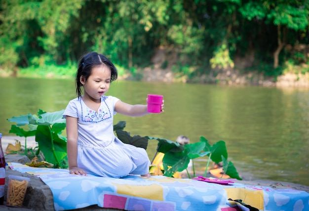 Niña sonriente sentada al aire libre en una roca grande y bebiendo cerca del río