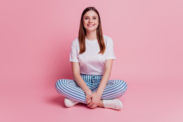 Niña sonriente sentada en el piso con las piernas cruzadas mira a la cámara sobre fondo rosa