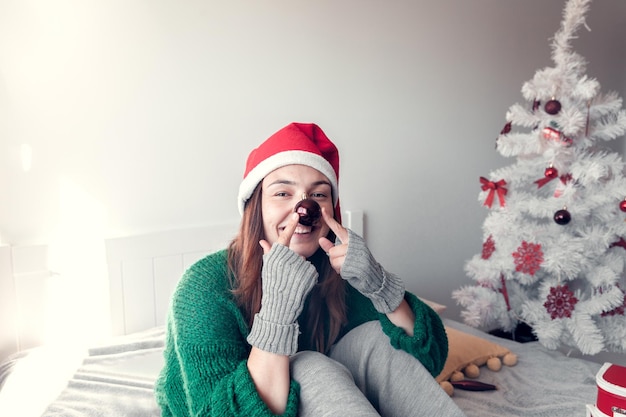 Niña sonriente se sienta en una cama contra el fondo de un árbol de Navidad y sostiene un juguete como nariz de payaso