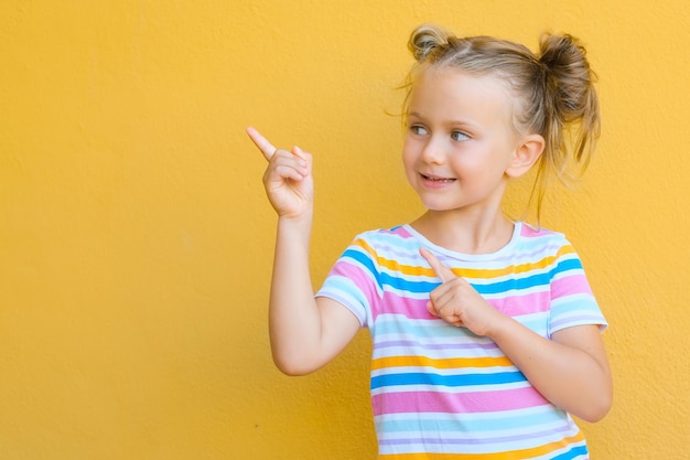 La niña sonriente se siente atraída por la atención que señala con el dedo el espacio de la copia El concepto de publicidad de productos y servicios Aislado en el fondo amarillo