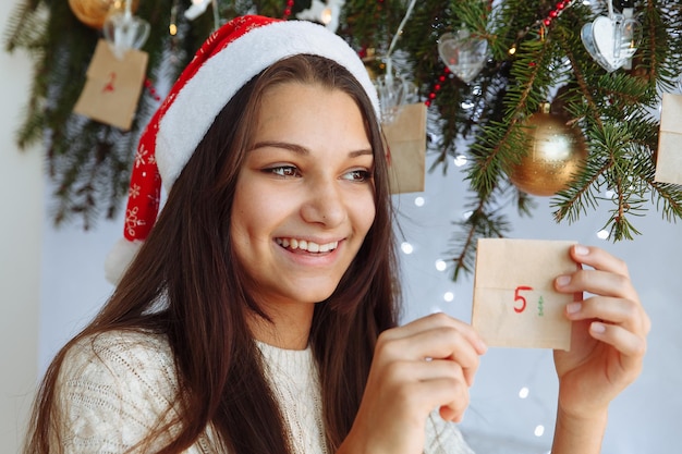Una niña sonriente con un sombrero de Navidad con un calendario de Adviento bricolaje en sus manos cerca del árbol de Navidad