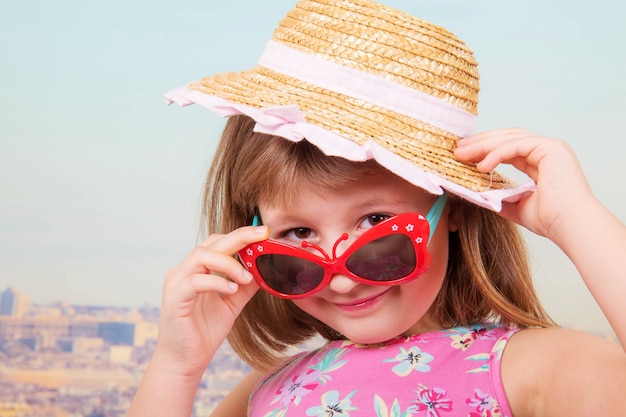 Foto niña sonriente con sombrero de paja y gafas de sol con parís como fondo
