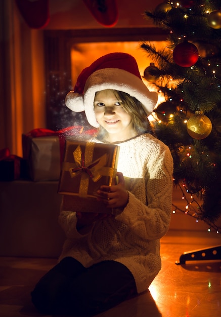 Niña sonriente sosteniendo brillante presente bajo el árbol de Navidad