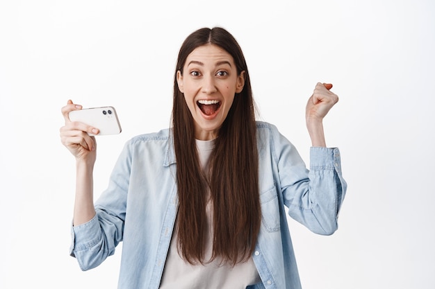 Niña sonriente sosteniendo el teléfono inteligente en posición horizontal, ganando en videojuegos, viendo videos en línea y celebrando la victoria, buenas noticias, de pie sobre una pared blanca