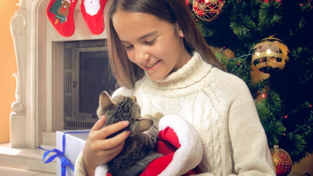 Niña sonriente en suéter blanco con lindo gatito junto al árbol de Navidad decorado