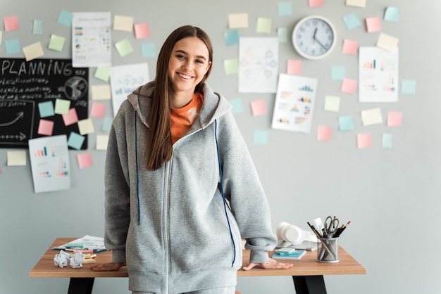 Foto niña sonriente con un suéter con capucha gris se inclinó contra un escritorio en su oficina.