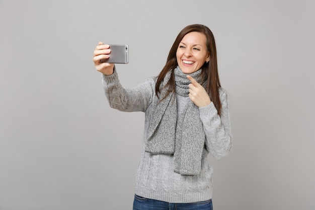 Niña sonriente en suéter gris bufanda apuntando con el dedo índice haciendo selfie en teléfono móvil haciendo videollamadas aisladas sobre fondo gris. Concepto de estación fría de emociones de estilo de vida de moda saludable.