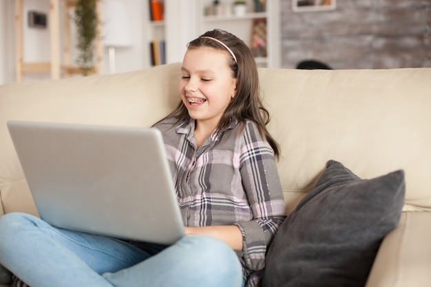 Niña sonriente con tirantes usando su computadora portátil en la sala de estar. Niño alegre.