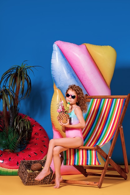 Niña sonriente en traje de baño y gafas de sol sentado en la silla de cubierta del arco iris con cóctel de piña con pajitas de colores y mostrando el pulgar hacia arriba y tomar el sol