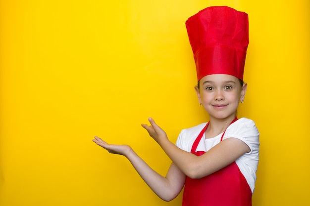 Foto niña sonriente en traje de chef rojo apunta con ambas manos a un copyspace