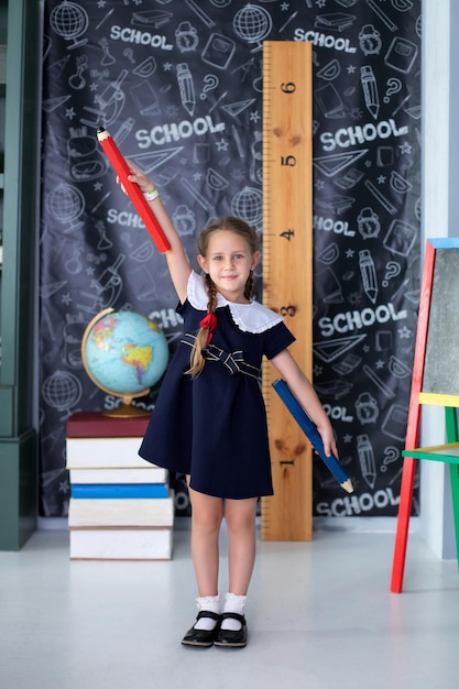 Una niña sonriente con uniforme escolar se para con grandes lápices en las manos en la escuela