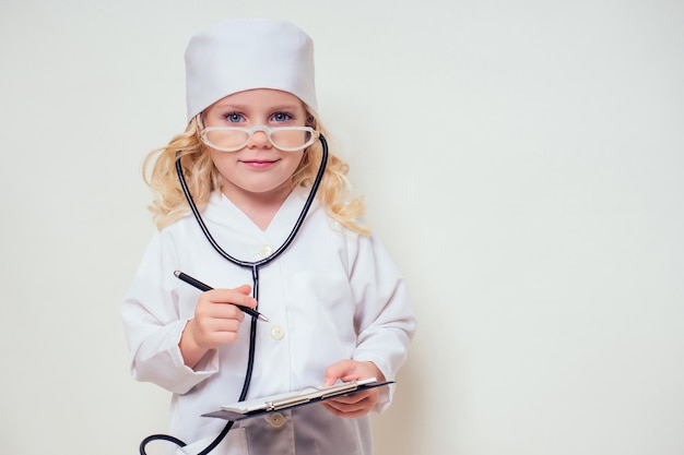 Niña sonriente en uniforme médico con estetoscopio de herramientas médicas escribiendo algo en el portapapeles sobre fondo blanco en el espacio de copia de estudio.