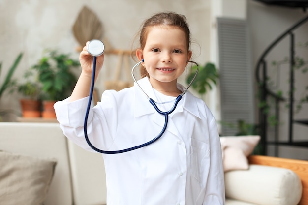 Niña sonriente en uniforme médico jugando con estetoscopio en casa.