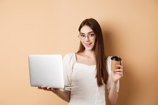 Niña sonriente en vasos trabajando en equipo portátil y tomando café para llevar. Freelancer usando computadora en el trabajo, de pie sobre fondo beige.