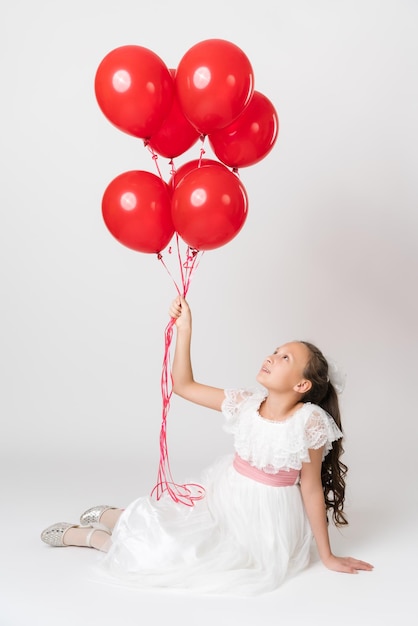 Niña sonriente vestida con un largo vestido blanco sosteniendo muchos globos rojos de fiesta en la mano mirando hacia arriba