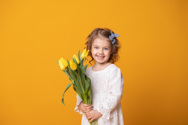 Niña sonriente en vestido blanco sobre fondo amarillo. Niño feliz alegre con ramo de flores de tulipanes.