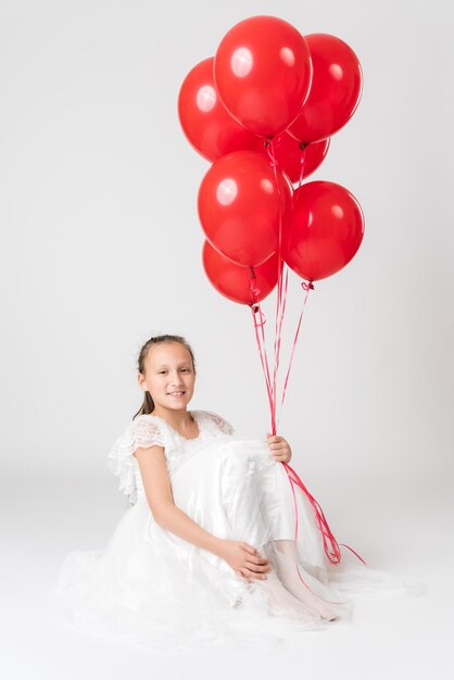 Niña sonriente con vestido blanco sosteniendo muchos globos rojos en la mano mirando a la cámara con emoción feliz