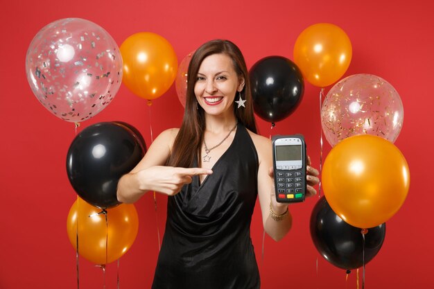 Niña sonriente en vestido negro con terminal de pago bancario moderno inalámbrico para procesar, adquirir pagos con tarjeta de crédito en globos de aire de fondo rojo. Concepto de fiesta de vacaciones de maqueta de cumpleaños de feliz año nuevo.