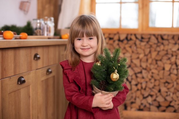 Niña sonriente en vestido rojo mostrando decorado pequeño abeto de Navidad en una olla en casa