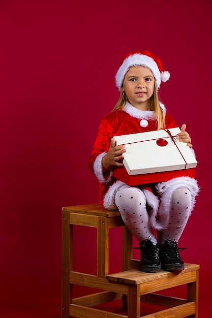 Niña sonriente, vestido con traje de Santa sentado en la silla de madera abierta regalo de Navidad, cuadro rojo-blanco. Pared aislada roja.