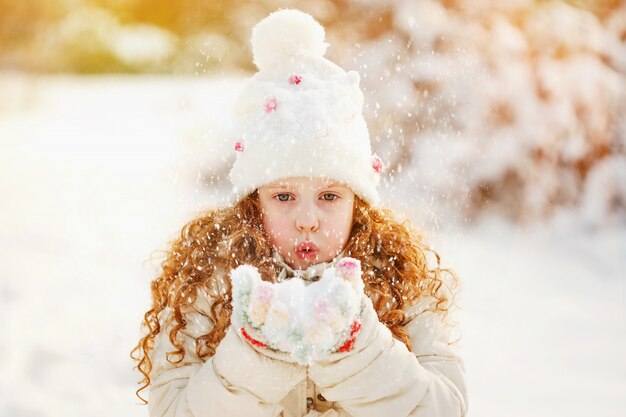 La niña sopla nieve con las manoplas, en un fondo del bokeh de los copos de nieve.