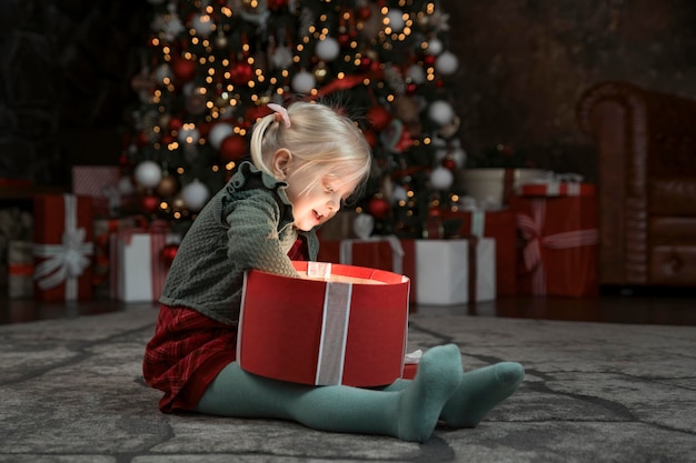Niña sorprendida abriendo regalo de Navidad cerca del árbol de Navidad Presente deseado Linda chica rubia con caja de regalo