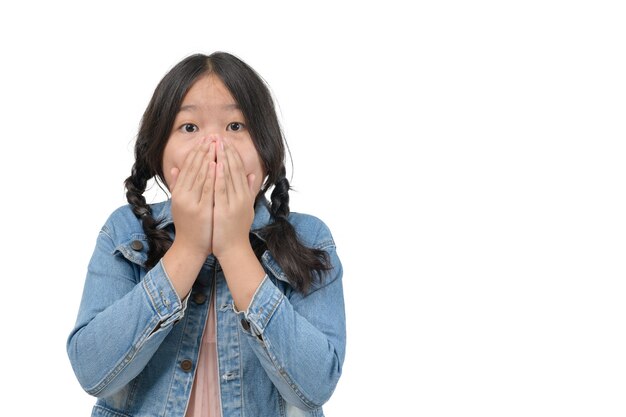 Niña sorprendida con chaqueta de mezclilla tapándose la boca y mirando a cámara aislada sobre fondo blanco, concepto de emoción conmocionada