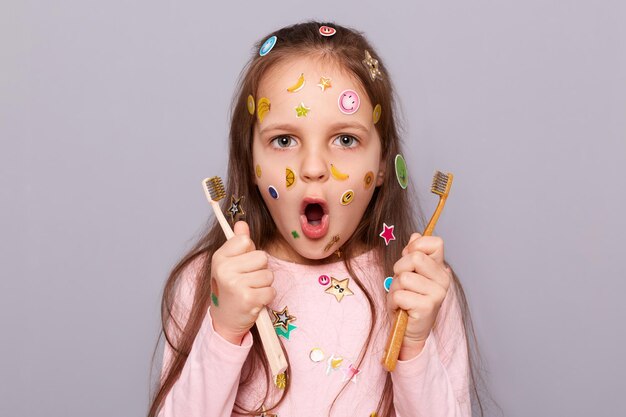 Niña sorprendida cubierta con pegatinas coloridas que sostienen dos cepillos de dientes mirando a la cámara con una mirada asombrada sorprendida por el cuidado oral usando una camisa rosa de pie aislada sobre un fondo gris