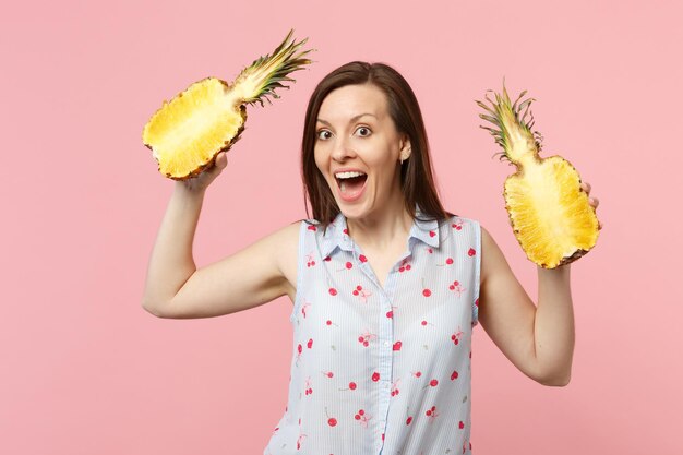 Niña sorprendida con ropa de verano que mantiene la boca abierta sostiene la mitad de la fruta fresca de piña madura aislada en un fondo rosa pastel. El estilo de vida vívido de la gente relaja el concepto de vacaciones. Simulacros de espacio de copia.