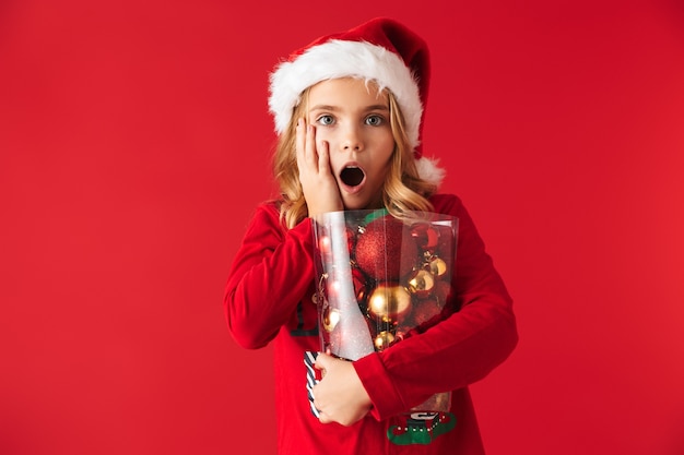 Niña sorprendida vistiendo traje de Navidad que se encuentran aisladas, sosteniendo un conjunto de juguetes de árbol de Navidad