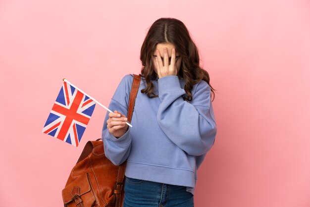 Niña sosteniendo una bandera del Reino Unido aislada en la pared rosa con expresión cansada y enferma