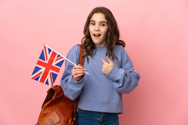 Niña sosteniendo una bandera del Reino Unido aislada en la pared rosa con expresión facial sorpresa