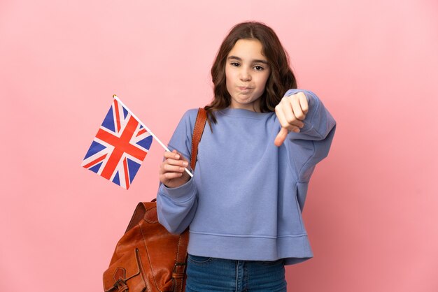 Niña sosteniendo una bandera del Reino Unido aislada sobre fondo rosa mostrando el pulgar hacia abajo con expresión negativa