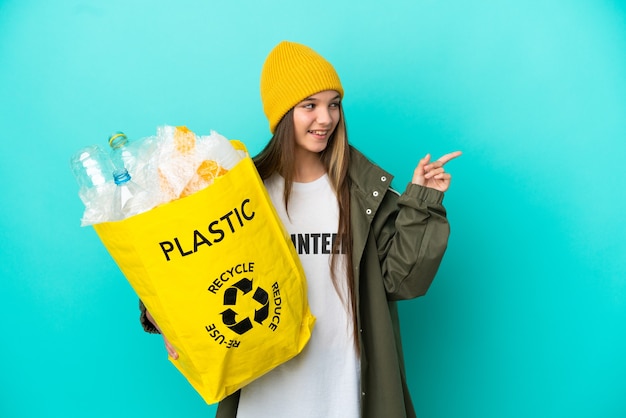 Foto niña sosteniendo una bolsa llena de botellas de plástico para reciclar sobre fondo azul aislado apuntando con el dedo hacia un lado y presentando un producto