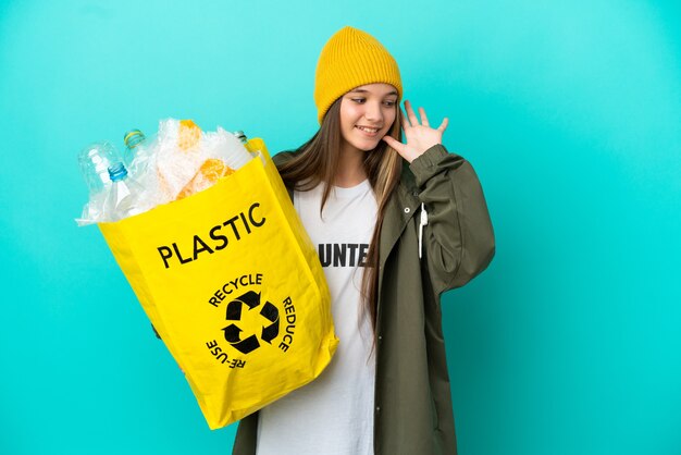 Niña sosteniendo una bolsa llena de botellas de plástico para reciclar sobre fondo azul aislado escuchando algo poniendo la mano en la oreja