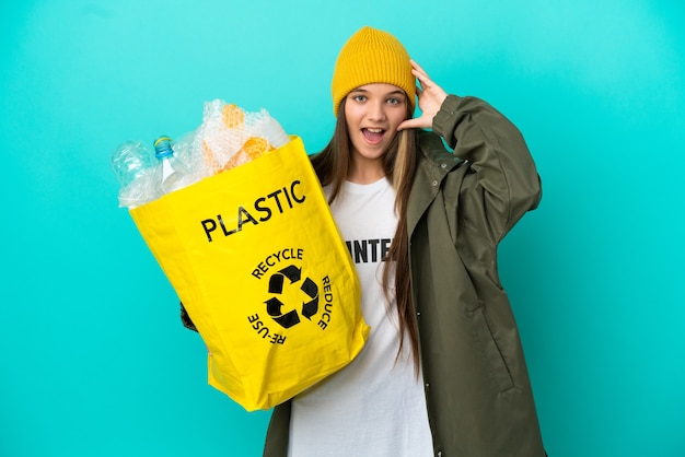 Niña sosteniendo una bolsa llena de botellas de plástico para reciclar sobre fondo azul aislado con expresión de sorpresa