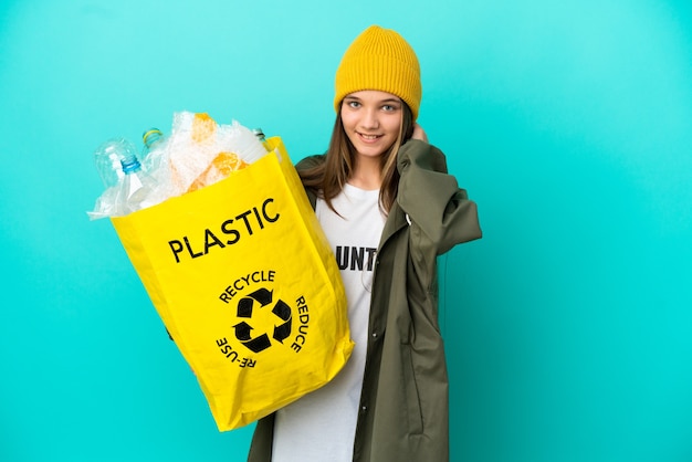 Niña sosteniendo una bolsa llena de botellas de plástico para reciclar sobre fondo azul aislado riendo