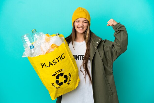 Niña sosteniendo una bolsa llena de botellas de plástico para reciclar sobre una superficie azul aislada haciendo un gesto fuerte