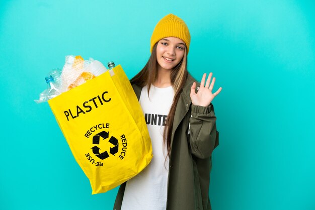 Niña sosteniendo una bolsa llena de botellas de plástico para reciclar sobre una superficie azul aislada saludando con la mano con expresión feliz