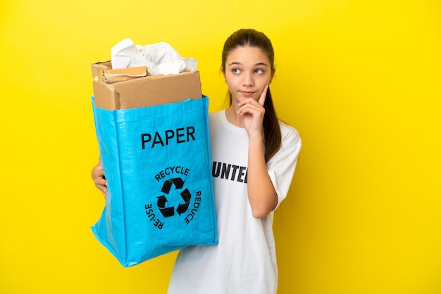 Niña sosteniendo una bolsa de reciclaje llena de papel para reciclar sobre fondo amarillo aislado pensando en una idea mientras mira hacia arriba