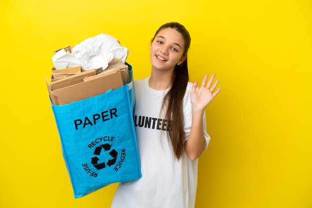 Niña sosteniendo una bolsa de reciclaje llena de papel para reciclar sobre fondo amarillo aislado saludando con la mano con expresión feliz