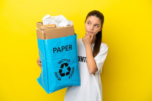 Niña sosteniendo una bolsa de reciclaje llena de papel para reciclar sobre pared amarilla aislada que tiene dudas