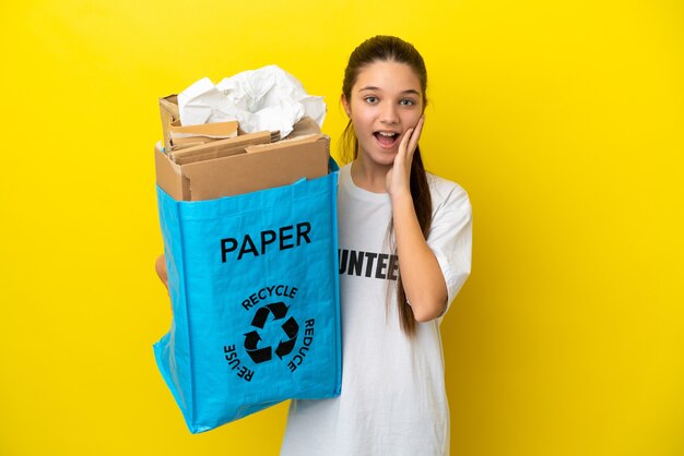 Niña sosteniendo una bolsa de reciclaje llena de papel para reciclar sobre una pared amarilla aislada con sorpresa y expresión facial conmocionada